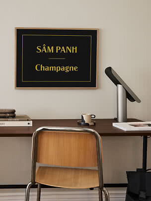 Framed black poster with gold text "SÂM PANH Champagne" above a desk and chair