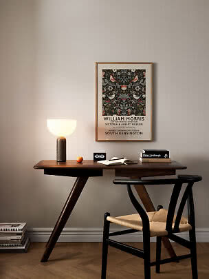 Træskrivebord med glødelampe, stol, bøger og indrammet William Morris-plakat på væggen
