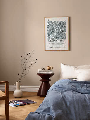 Soveværelse med blå sengetæppe, sengebord og indrammet William Morris-plakat på beige væg