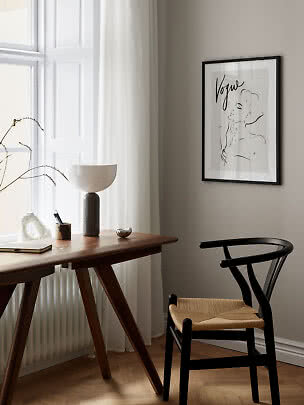 Minimalist workspace with wooden desk, black chair, table lamp, and framed Vogue line art print.