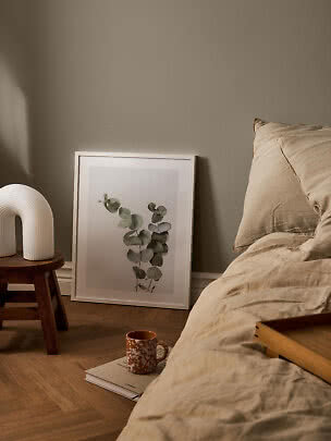 Cozy bedroom with beige bedding, botanical print, white lamp on stool, and mug on a book
