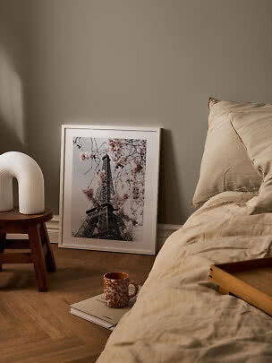 Gemütliches Schlafzimmer mit beiger Bettwäsche, Tischlampe und gerahmtem Eiffelturm-Poster am Boden