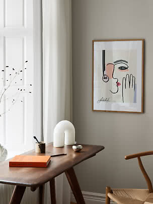 Gerahmtes abstraktes Gesichtsposter an beiger Wand über Holztisch in hellem, minimalistischem Raum