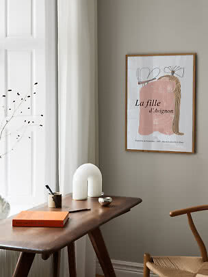 Framed art print above a wooden desk in a bright corner with book, vase and chair