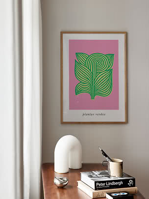 Oprawiony plakat z zieloną ilustracją botaniczną na różowym tle nad drewnianym stołem