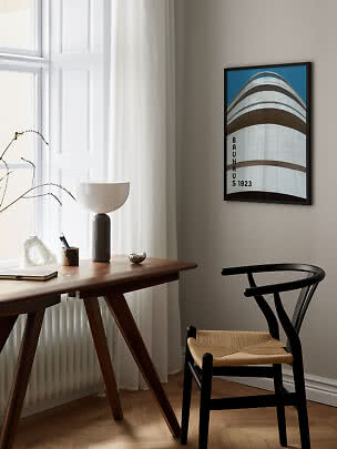 Oprawiony plakat Bauhaus 1923 na ścianie nad biurkiem i czarnym krzesłem w jasnym pokoju