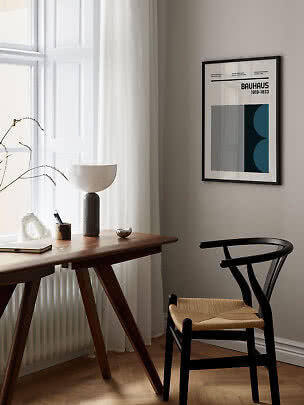 Bauhaus-plakat i sort ramme på lys væg over skrivebord og sort stol ved vindue