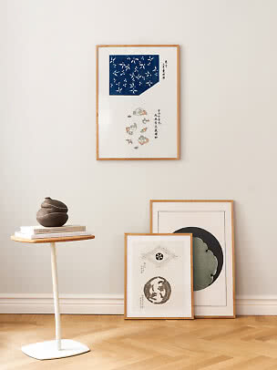 Træindrammede kunsttryk på lys væg, med lille sidebord, bøger og mørk keramisk vase.