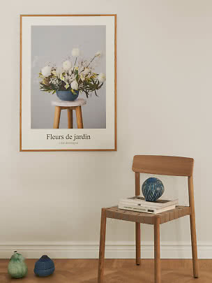 Inramad blomsterposter på beige vägg ovanför trästol med böcker och blå vas
