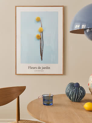 Póster botánico enmarcado con flores amarillas esféricas sobre fondo azul claro sobre mesa de madera