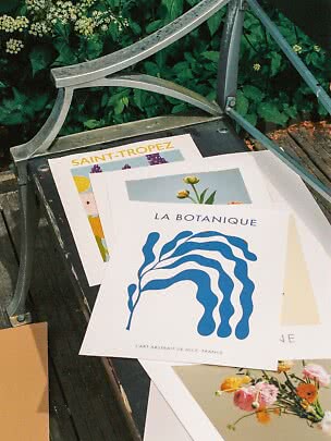 Kunstprint og blomsterplakater ligger på en bænk i haven under metalarmstøtte.