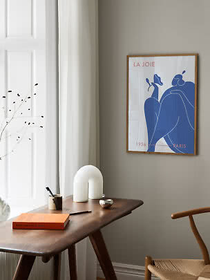 Træindrammet kunstplakat med blå figur på beige væg over et træbord ved et vindue