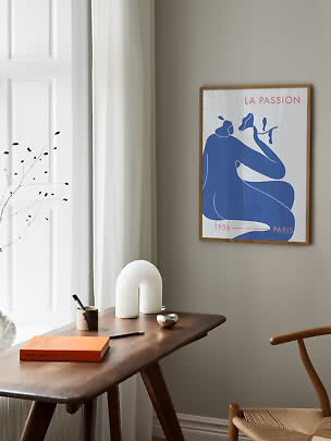 Framed art print with blue abstract figure above a wooden desk in a bright room corner