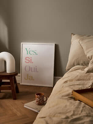 Poster con la scritta "Yes. Si. Oui. Ja." appoggiato al muro accanto al letto in una camera calda