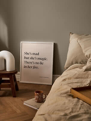 Poster con citazione in cornice appoggiato al muro vicino al letto, sgabello, lampada e tazza