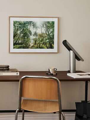 Stampa incorniciata di palme sopra scrivania con sedia, lampada, tazza e libri