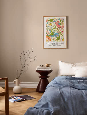 Framed William Morris floral poster above a bed in a beige bedroom with blue duvet