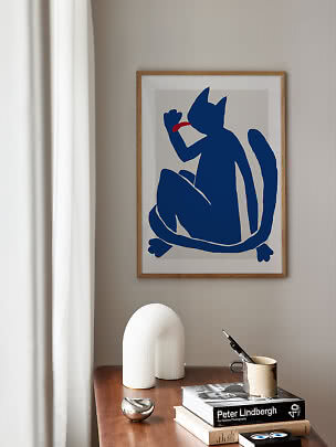 Indrammet plakat med blå kat og rød tunge på beige væg over et træbord.