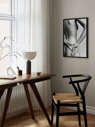 Minimalistische werkplek met houten bureau, zwarte stoel, tafellamp en zwart-wit poster aan de muur.