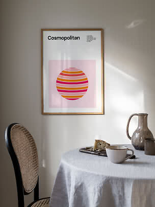 Gerahmtes Cosmopolitan-Poster mit rosa-orangefarbenem Grafikmotiv an heller Wand über Esstisch.