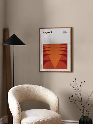 Ingelijste Negroni-poster met oranje grafisch motief aan beige muur boven witte fauteuil
