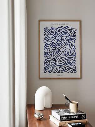 Framed art print with blue wave pattern above a wooden table with books and a white lamp