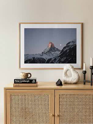 Gerahmtes Bergmotiv mit schneebedecktem Gipfel und Mond über einem Holzsideboard