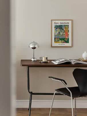 Schreibtisch mit Chromlampe, Becher, offenem Buch und gerahmtem Paul-Gauguin-Poster an beiger Wand