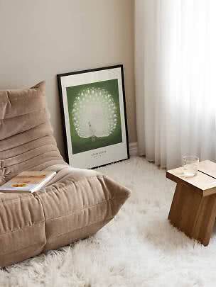Framed peacock print leaning against the wall beside a beige lounge chair and wooden side table