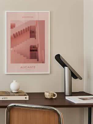 Inramad Alicante-poster med rosa trappa ovanför skrivbord med lampa, böcker, vas och kopp