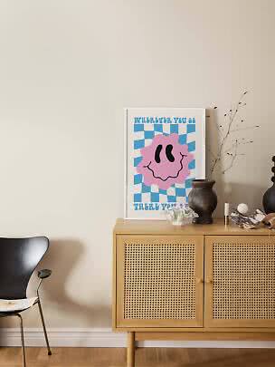 Poster colorato con smiley rosa su sfondo blu a scacchi, appoggiato su mobile in legno con vaso.