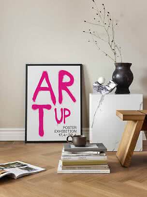Inramad konstposter med rosa texten ”ART UP” lutad mot vägg på trägolv
