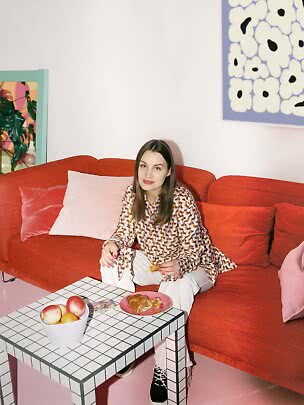 Woman sitting on a red sofa with a croissant by a tiled coffee table with fruit in a colorful room