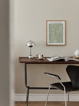 Minimalistisk skrivebord med kromlampe, krus, åben bog og botanisk plakat på beige væg
