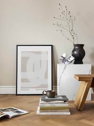 Poster astratto con cornice nera appoggiato al muro, accanto a libri, vaso e piedistallo in marmo