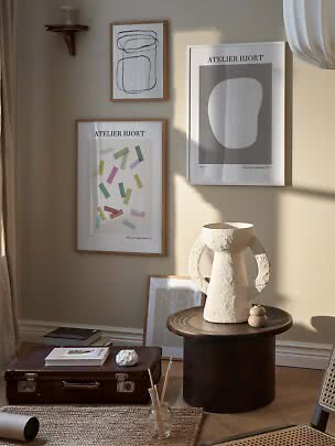 Mur avec trois affiches abstraites au-dessus d’une table ronde avec grand vase texturé au soleil
