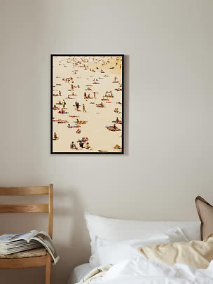 Gerahmter Stranddruck mit Sonnenbadenden über einem Bett in einem hellen Schlafzimmer