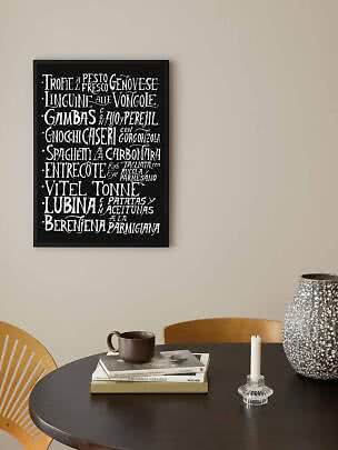Black framed poster with white handwritten menu text above a round table with mug and vase