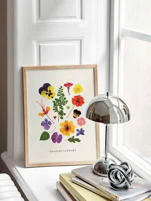 Indrammet blomsterplakat med farverige pressede blomster på en hvid vindueskarm ved siden af en kromlampe