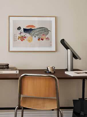 Inramat konsttryck med frukt och grönsaker ovanför ett mörkt skrivbord med lampa och stol