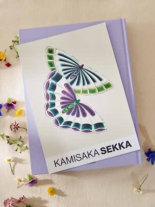 Karte mit farbiger Schmetterlingsillustration auf lila Buch, umgeben von kleinen Blumen