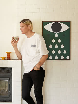 Person in weißem T-Shirt und schwarzer Hose hält ein Glas vor weißer Ziegelwand mit Grafikposter.