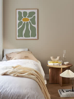 Schlafzimmer mit Bett, rundem Holztisch und abstraktem Blumenposter an beiger Wand