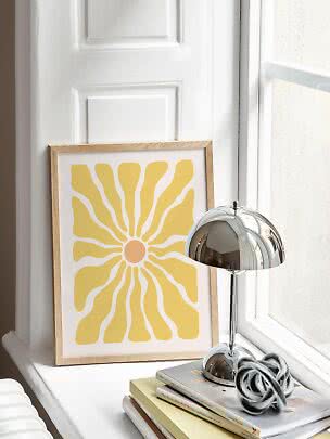 Framed yellow sun print on a windowsill beside a chrome table lamp and stacked books