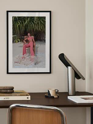 Framed art print of woman in patterned outfit on chair by water and tropical plants