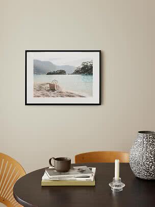 Framed beach print with basket by clear water above a dark table with vase, candle and mug