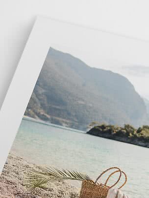 Large framed beach print leaning against a wall with a woven bag and palm leaf in front