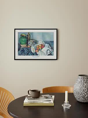 Indrammet Cézanne-plakat med stilleben hænger over rundt bord med bøger, krus, lysestage og stor vase