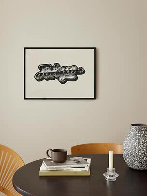Ingelijste Tokyo-typografieposter aan beige muur boven donkere tafel met boeken, mok, kandelaar en vaas