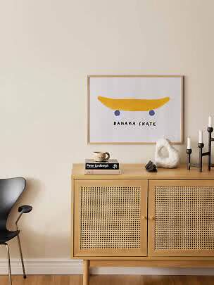 Gerahmtes Poster mit Banane als Skateboard über hellem Holzschrank mit Geflechttüren